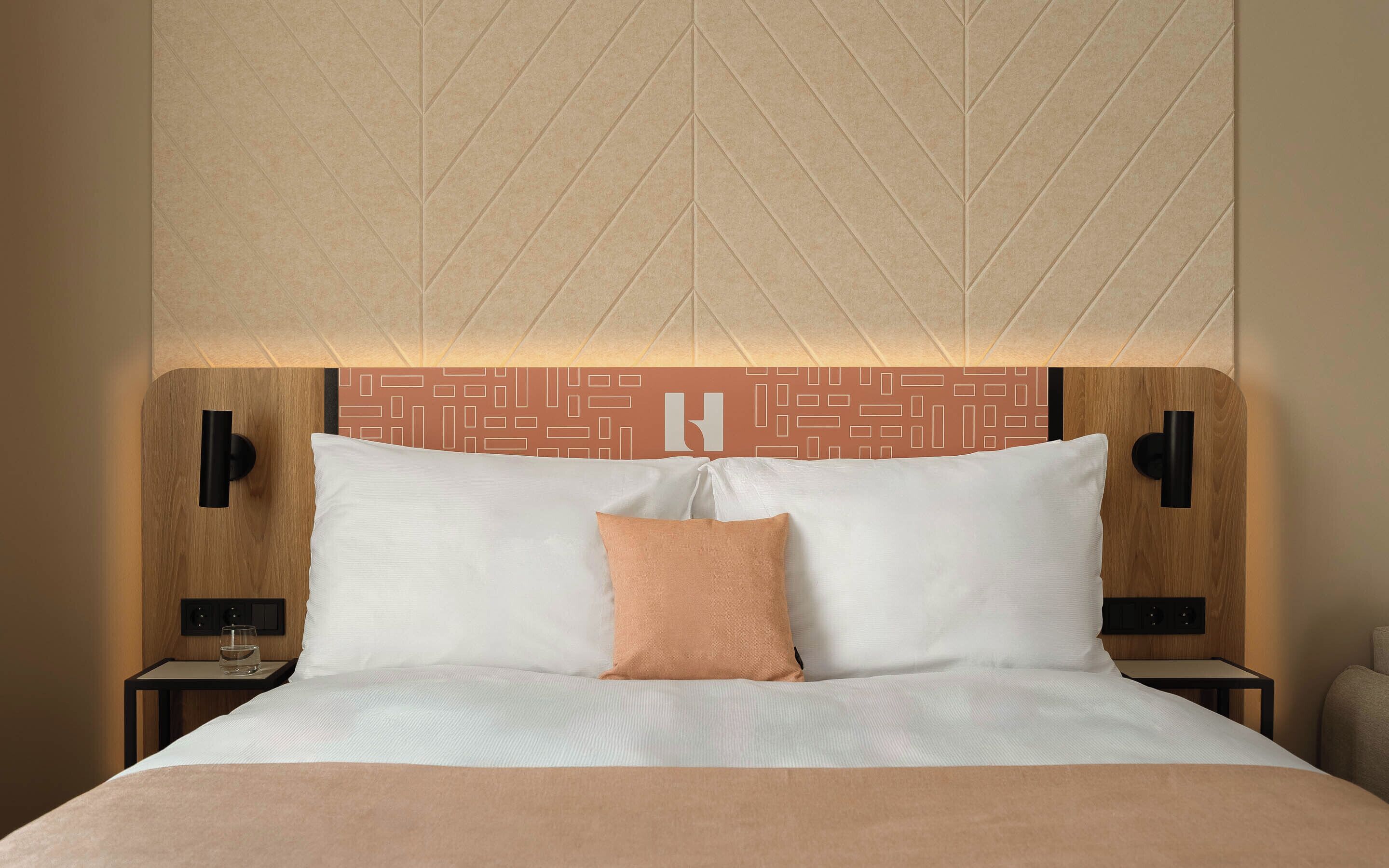 A neatly made bed with white and peach pillows, a wooden headboard with a geometric pattern, and two bedside tables with black lamps.