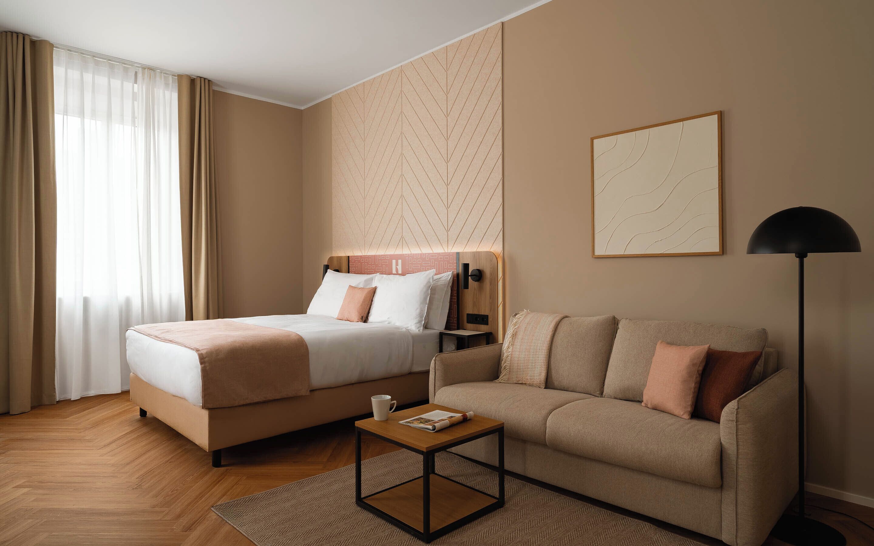 A hotel room with a bed, a sofa, a small table, a lamp, and a wall with a geometric pattern.