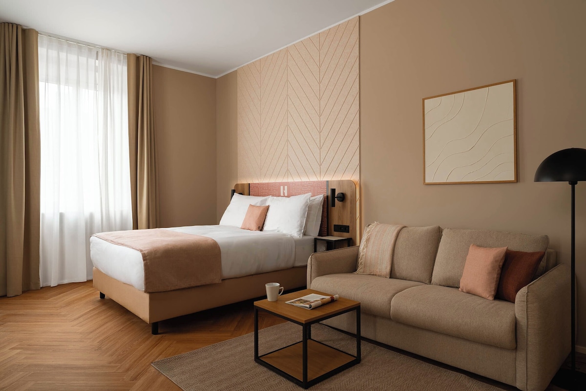 A hotel room with a bed, a sofa, a small table, a lamp, and a wall with a geometric pattern.