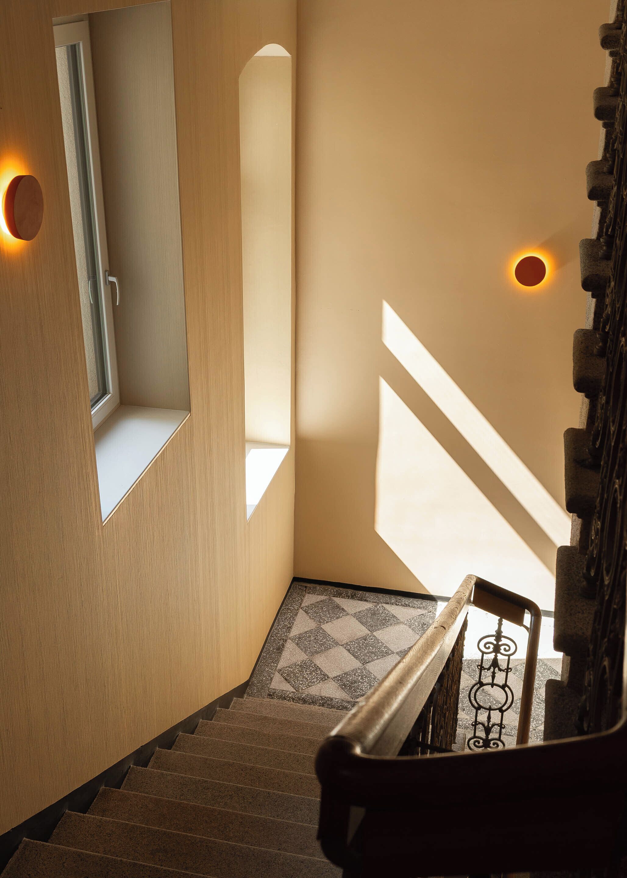 Un escalier recouvert d’une moquette à motifs, avec une main courante et un luminaire fixé au mur.