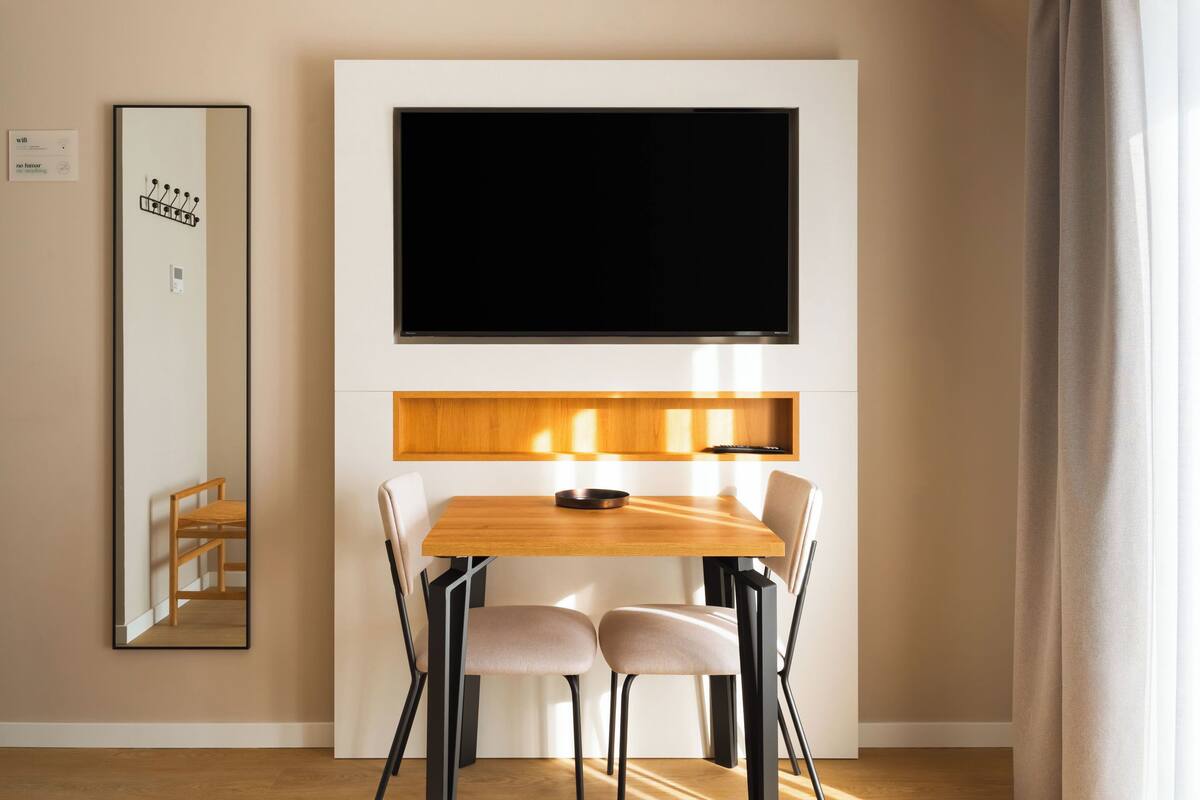 Un comedor moderno con una mesa de madera, cuatro sillas, un televisor de pantalla plana montado arriba y un espejo en la pared.