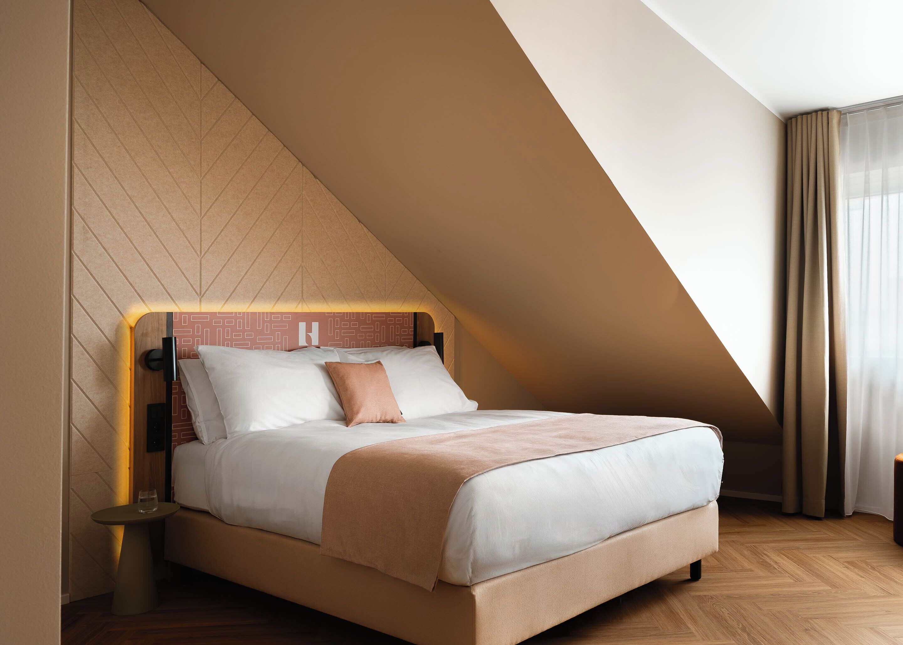 Ett modernt hotellrum med ett sluttande tak, en säng med vita och beige sängkläder, ett litet sängbord och ett trägolv.