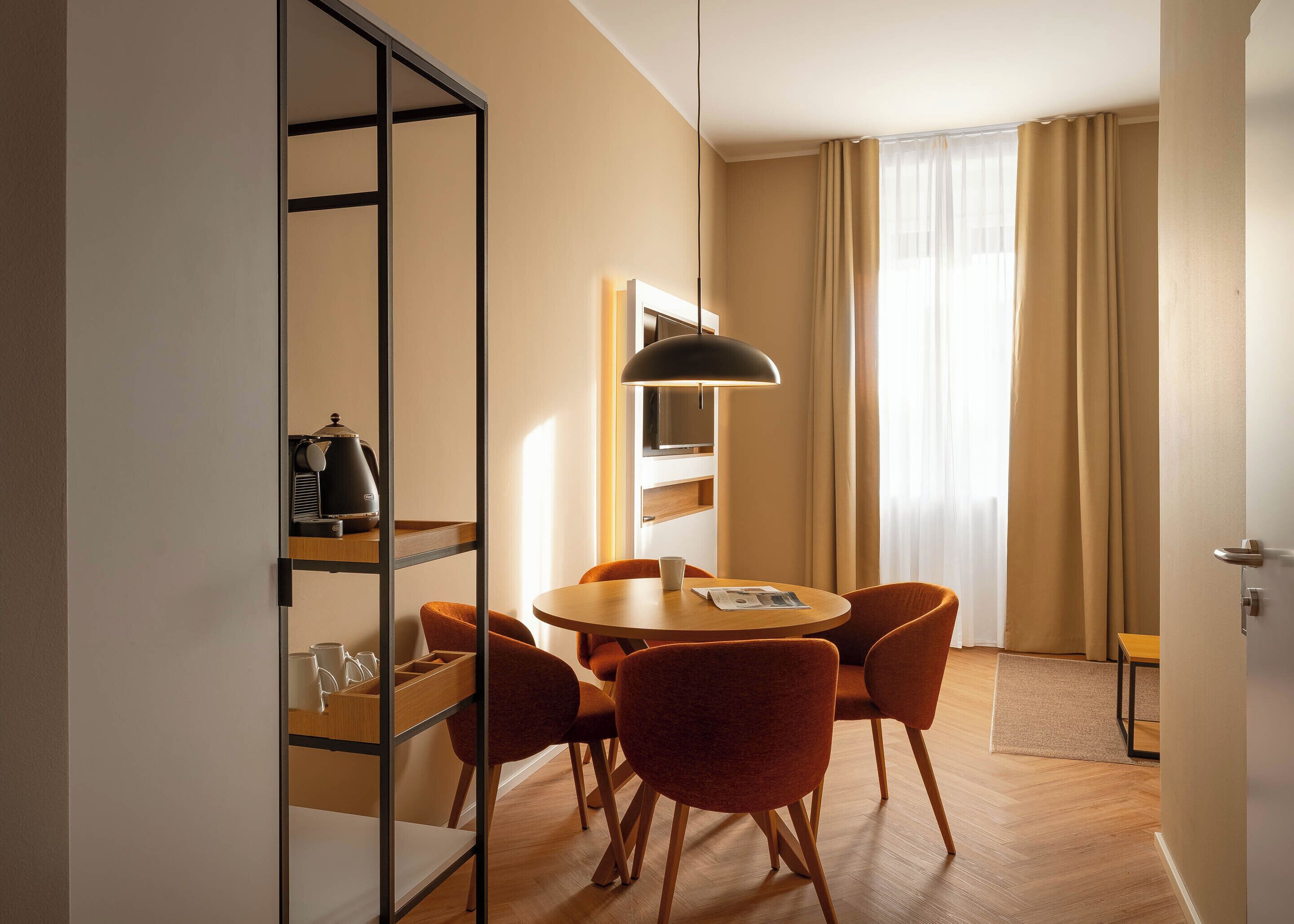 A hotel room with a round wooden table, four orange chairs, a black pendant light, a bookshelf, and a large window with curtains.