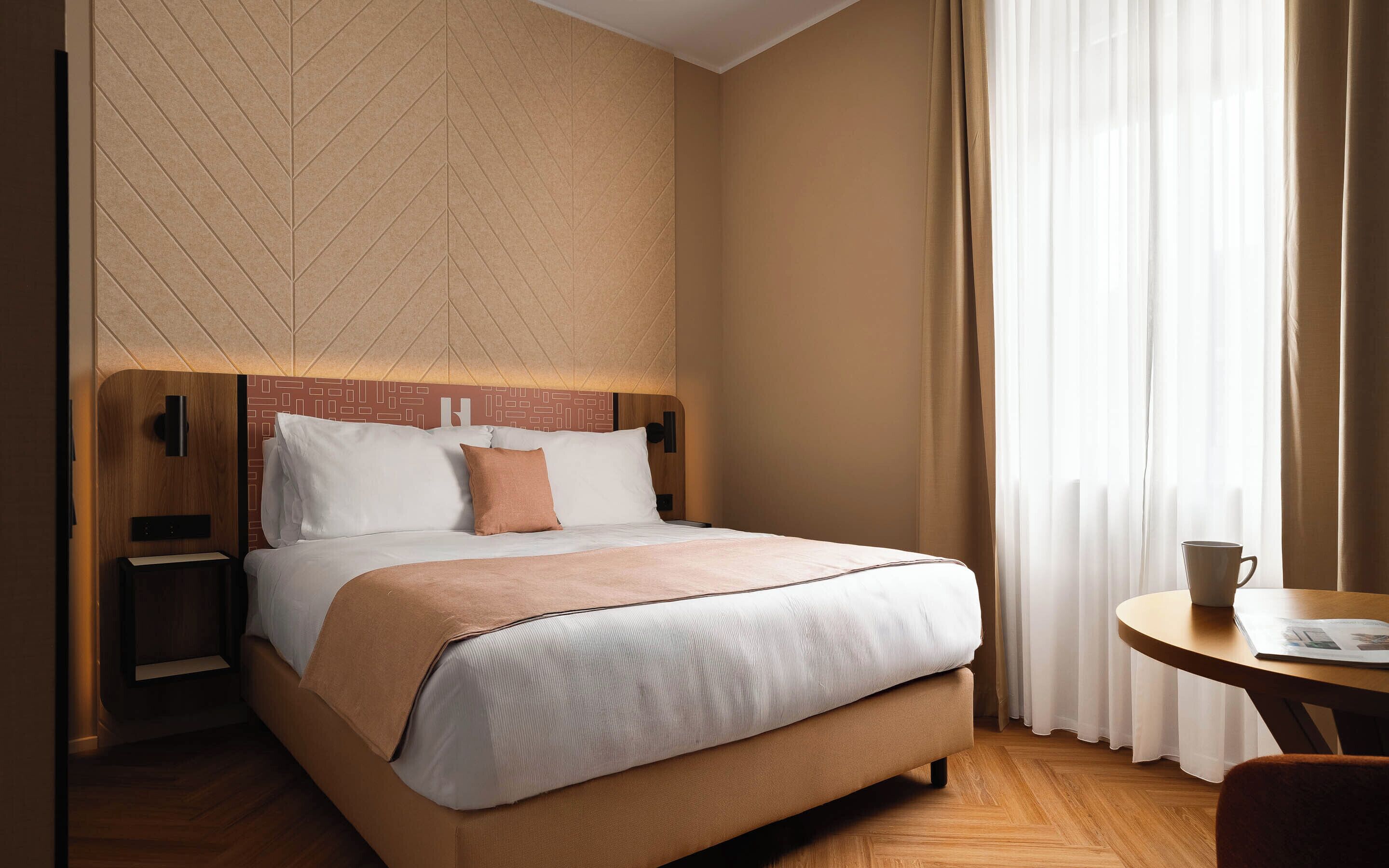 Habitación de hotel con cama, mesitas de noche, televisor montado en la pared y ventana con cortinas.