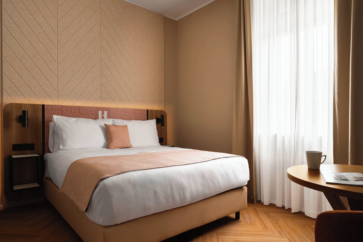 A hotel room with a bed, bedside tables, a wall-mounted TV, and a window with curtains.