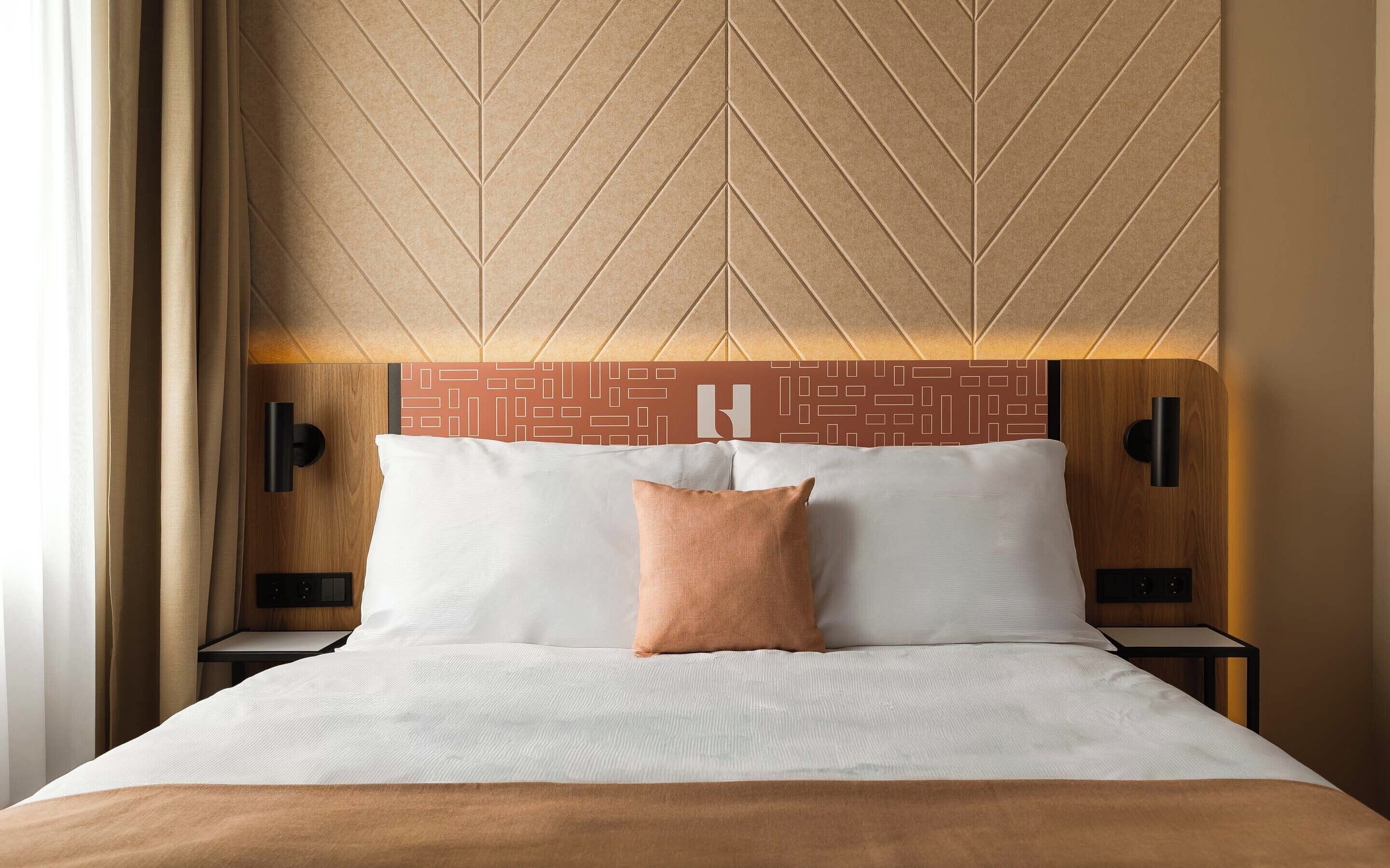 A hotel room with a bed, two bedside tables, a headboard with a geometric pattern, and a wall-mounted light fixture.