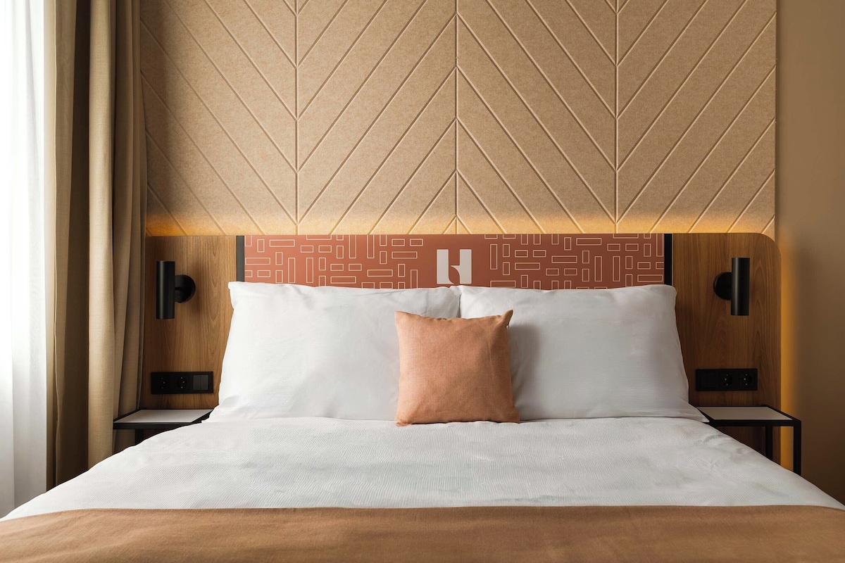 A hotel room with a bed, two bedside tables, a headboard with a geometric pattern, and a wall-mounted light fixture.