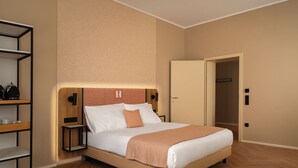 Habitación de hotel con cama, mesita de noche, estante con objetos y puerta que da a otra habitación.