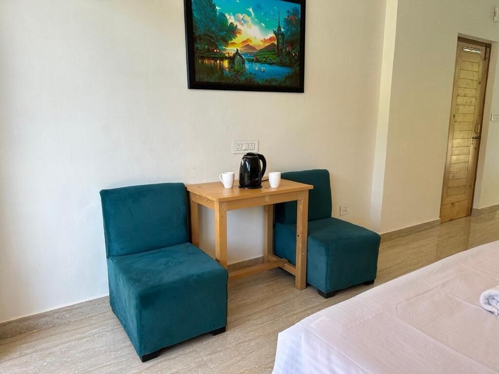 Una habitación de hotel con una cama, dos sillones azul verdosos, una pequeña mesa de madera con una tetera y un cuadro en la pared.