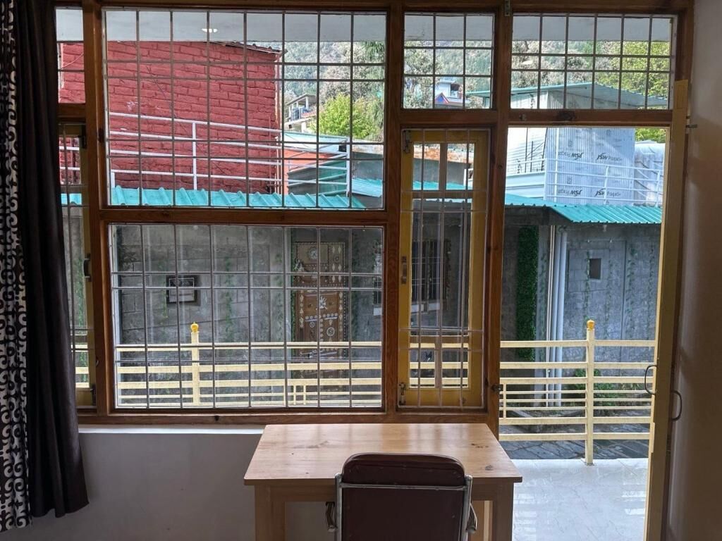 A room with a wooden table and a chair, a large window with metal bars, and a view of buildings outside.