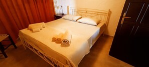 A bed with white linens and rolled towels, a bedside table with a lamp, and a door to the right.