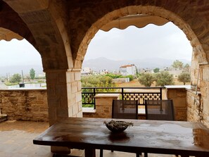 A stone balcony with a table and chairs, overlooking a cityscape with buildings and greenery.