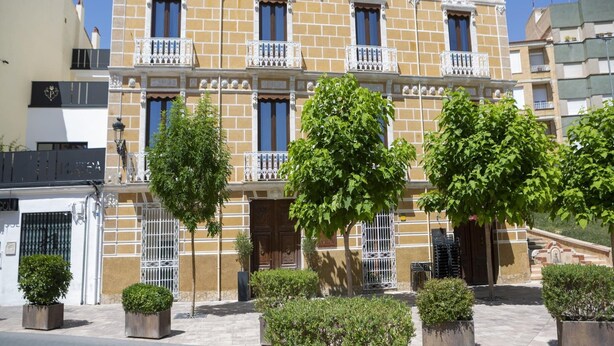 Un immeuble à plusieurs étages doté de balcons, d’une entrée fermée par un portail et de plantes en pot disposées au premier plan.