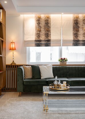A living room with a green sofa, a wooden bookshelf, a lamp, a window with sheer curtains, and a tray with decorative items. - Voula Calm Living | Igias 5-7 | Voula 166 73, Bill & John Villas (Vari - Voula - Vouliagmeni)