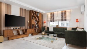 A modern living room with a flat-screen TV, wooden bookshelves, a green sofa, and a glass coffee table. - Voula Calm Living | Igias 5-7 | Voula 166 73, Bill & John Villas (Vari - Voula - Vouliagmeni)