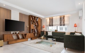 A modern living room with a flat-screen TV, wooden bookshelves, a green sofa, and a glass coffee table. - Voula Calm Living | Igias 5-7 | Voula 166 73, Bill & John Villas (Vari - Voula - Vouliagmeni)