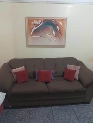 Ein braunes Sofa mit drei roten und zwei beigen Kissen vor einer schlichten Wand mit einem gerahmten abstrakten Gemälde.