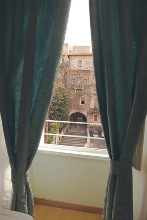 Blick durch ein Fenster mit Vorhängen, zeigt ein Gebäude mit einer steinernen Fassade und einer Treppe, die zu einem Eingang führt.