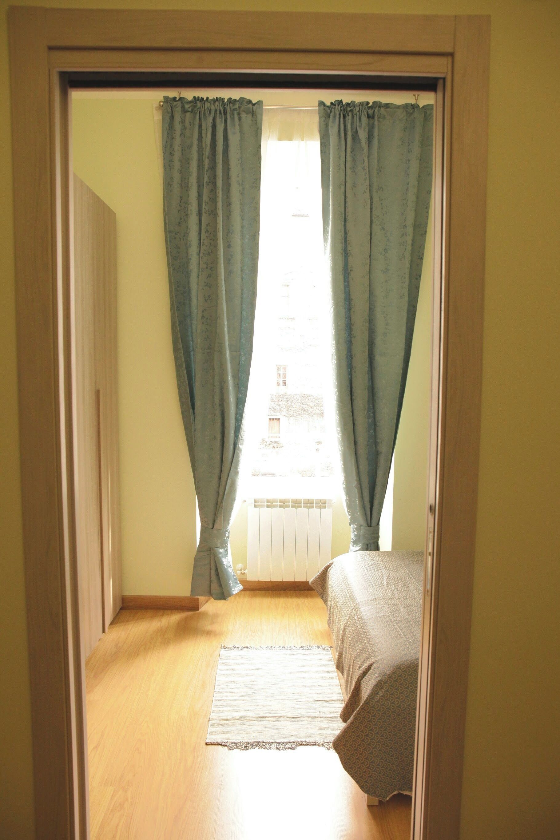 A room with a bed, a window with curtains, a radiator, and a wooden floor.