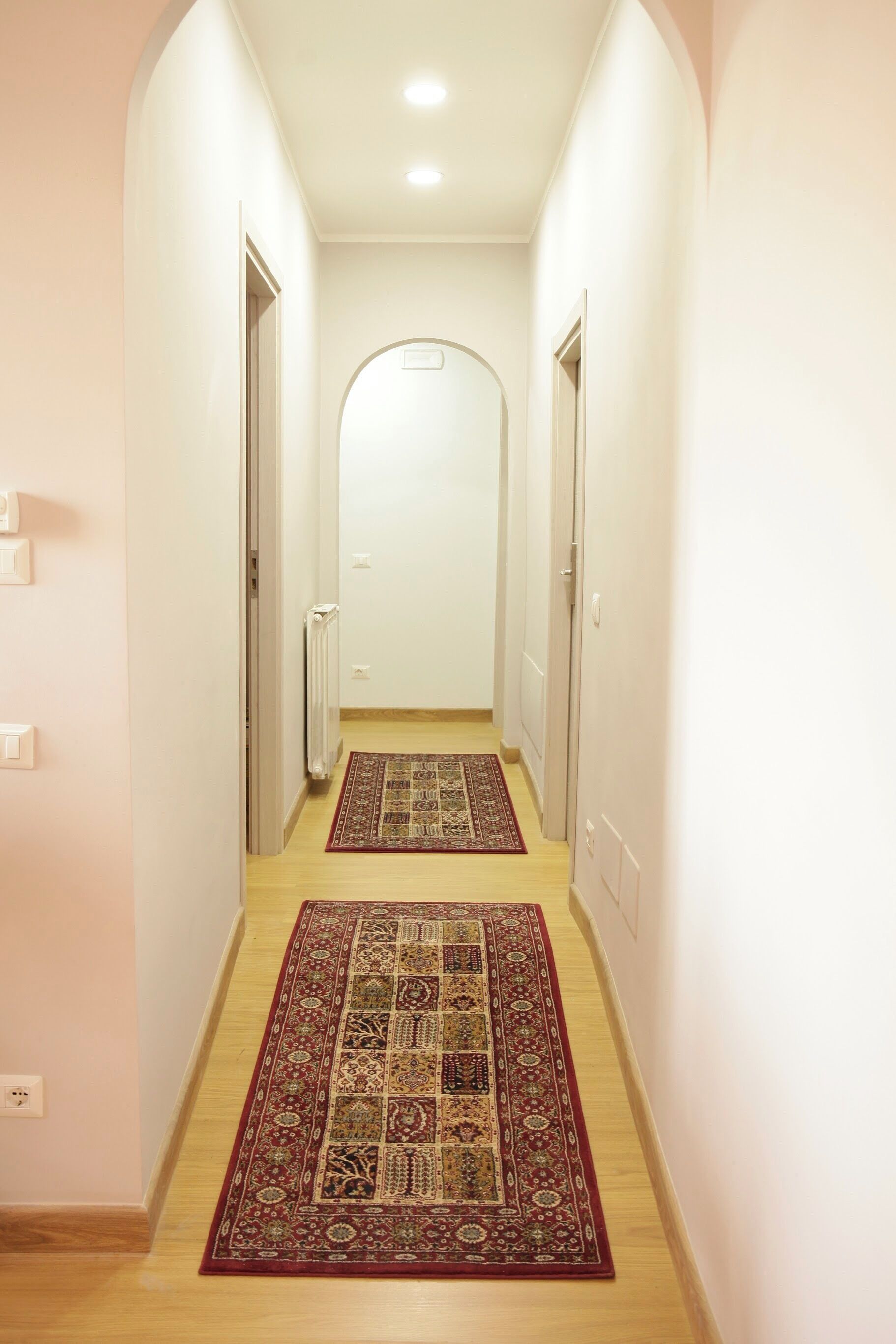 A narrow hallway with two doors on either side, a radiator on the left, and a patterned rug on the floor.