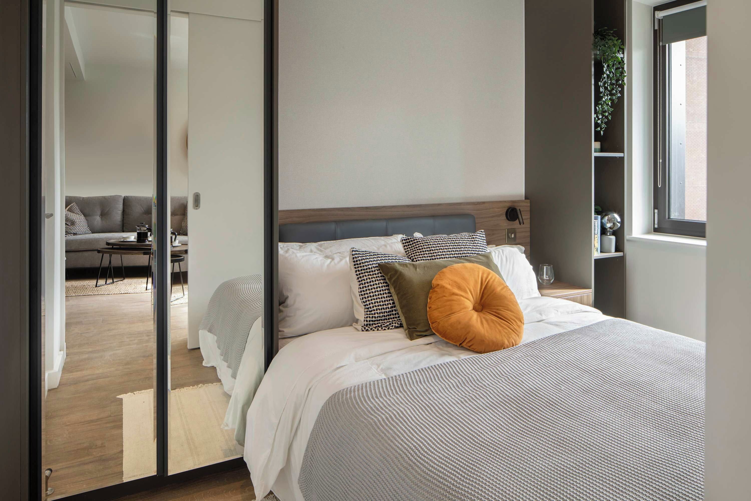 Une chambre moderne avec un grand lit, une tête de lit en bois et une vue sur l’espace de vie, accessible par une porte vitrée.