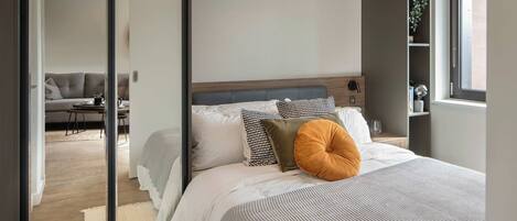 Une chambre moderne avec un grand lit, une tête de lit en bois et une vue sur l’espace de vie, accessible par une porte vitrée.