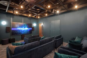 A modern home theater with dark sofas, a large screen, and ceiling lights. - One-bedroom | Gym, CoWorking, Library | StayCozy (Croydon)