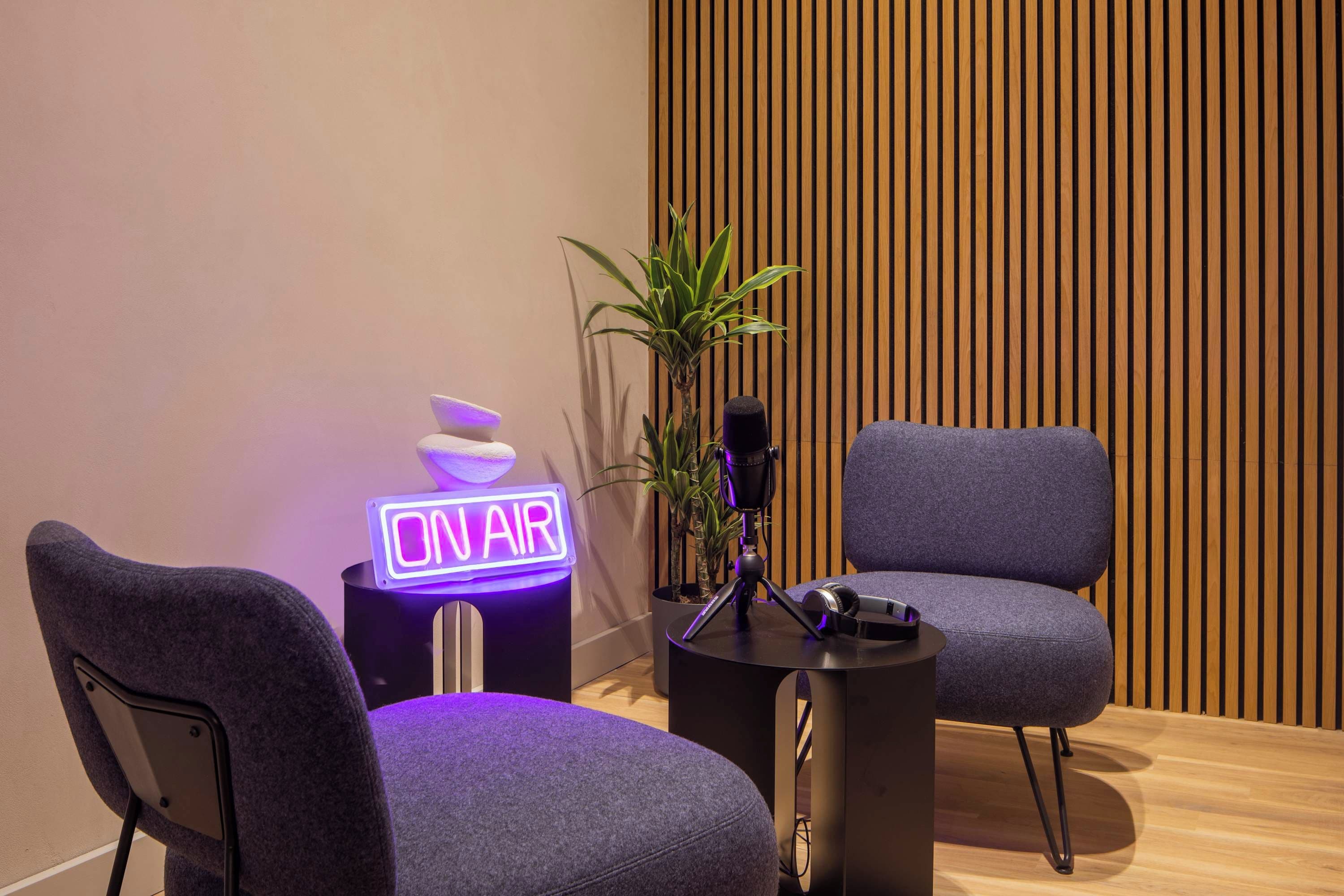 A modern room with a neon sign reading "ON AIR," a microphone on a stand, and two grey chairs.