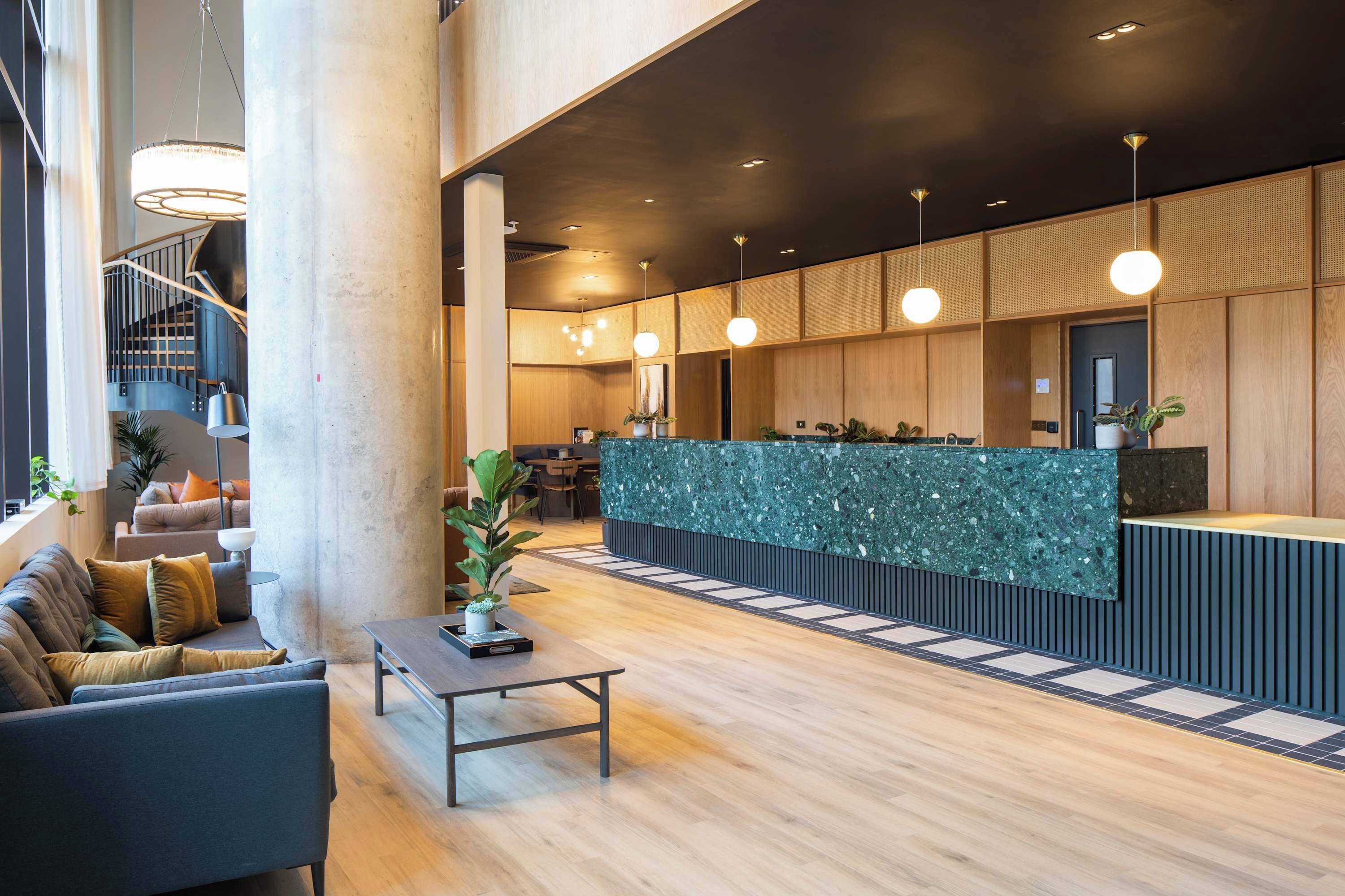 Een moderne lobby met een receptiebalie, een zithoek, een trap en potplanten.