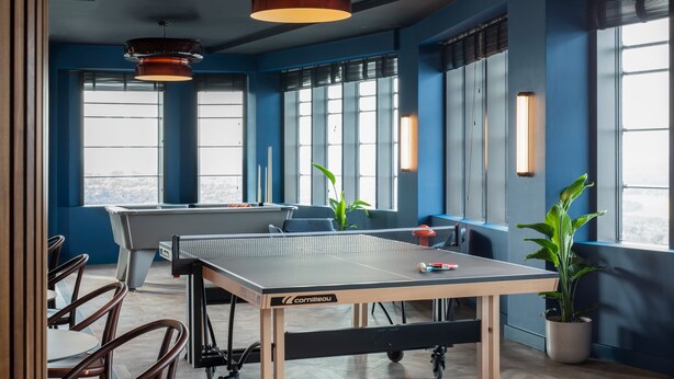Een tafeltennistafel in een kamer met grote ramen, stoelen en potplanten.