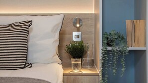 Een net opgemaakt bed met een gestreept kussen, een nachtkastje met een glas water, een kleine plant en een wandlamp.