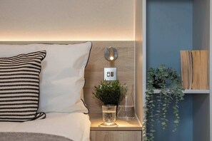 A neatly made bed with a striped pillow, a bedside table with a glass of water, a small plant, and a wall-mounted light fixture. - Roomy Studio | Workspace & Laundry Room | StayCozy (Croydon)