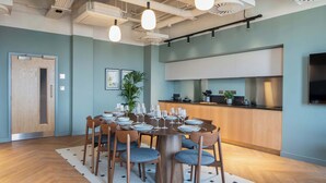 Een moderne eetruimte met een ronde tafel voor zes personen, omringd door houten stoelen met blauwe kussens. Op de achtergrond is een keuken zichtbaar met witte kasten en een zwart aanrecht.