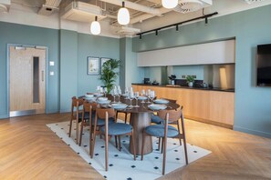 A modern dining area with a round table set for six, surrounded by wooden chairs with blue cushions. A kitchen with white cabinets and a black countertop is visible in the background. - Roomy Studio | Workspace & Laundry Room | StayCozy (Croydon)