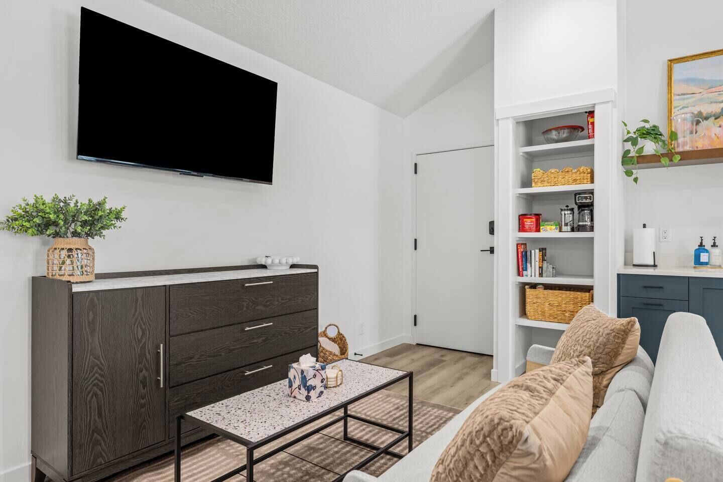 Una sala de estar moderna con un televisor de pantalla plana, un mueble de madera oscura, estantes blancos y un sofá con cojines estampados.