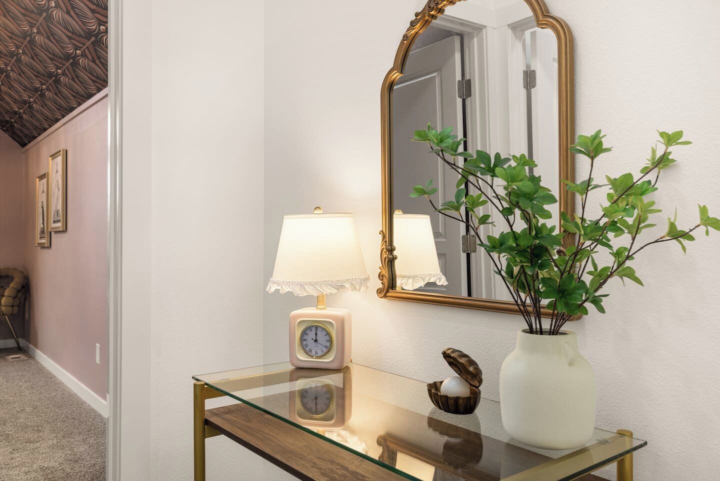 Un couloir avec une console en verre, un miroir fixé au mur, une horloge et une plante en pot.