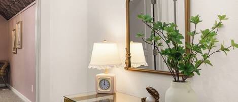 Un pasillo con una mesa consola de vidrio, un espejo montado en la pared, un reloj y una planta en maceta.