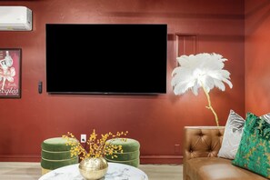 A living room with a large flat-screen TV, a green ottoman, a brown leather sofa, and a white feathered plant. - The High Roller House | Hot Tub & Pool Table (Meridian)