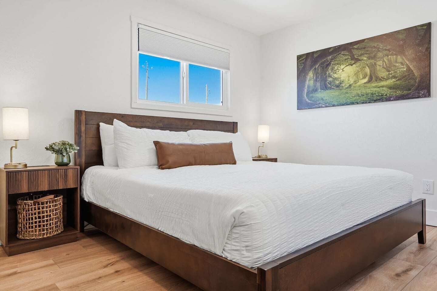 A neatly made bed with white linens and a brown accent pillow, flanked by two bedside tables with lamps, a window with blinds, and a forest-themed painting on the wall.