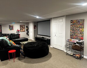 A living room with a black leather couch, a black bean bag, a red stool, a TV, and a wall with comic book posters. - Cozy Redford Getaway • Near Detroit & DTW Airport (Redford)