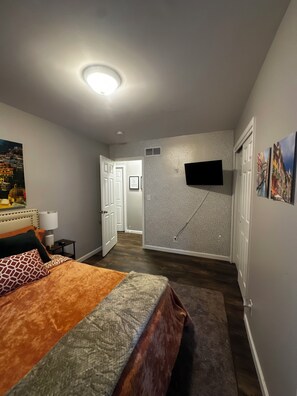 Un dormitorio con una cama, un televisor montado en la pared y una puerta que da a otra habitación.