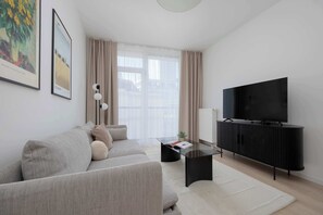 Ein modernes Wohnzimmer mit Sofa, Couchtisch, Fernseher auf einem Schrank und einem großen Fenster mit Vorhängen.