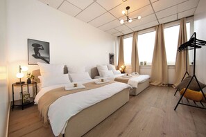 Ein Hotelzimmer mit zwei Betten, einem Nachttisch mit Lampe, einem Kleiderständer und einem großen Fenster mit Vorhängen.