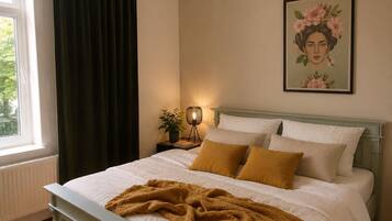 A bedroom with a bed, a nightstand, a lamp, a window with curtains, and a framed picture on the wall.