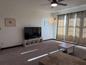 A living room with a flat-screen TV on a stand, a ceiling fan, and a large window with curtains.