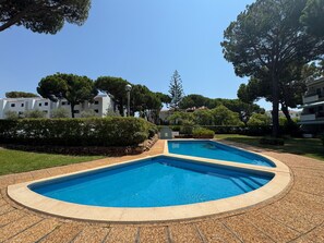 Uma área de piscina com duas piscinas, rodeada por um caminho pavimentado, árvores e edifícios ao fundo.