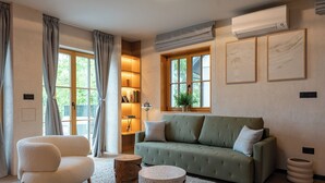 Ein Wohnzimmer mit einem grünen Sofa, einem weißen Sessel, einem runden Couchtisch und einem Holznachttisch. Zwei Fenster mit Vorhängen, ein Bücherregal und eine wandmontierte Klimaanlage sind ebenfalls vorhanden.