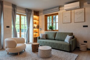 Ein Wohnzimmer mit einem grünen Sofa, einem weißen Sessel, einem runden Couchtisch und einem Holznachttisch. Zwei Fenster mit Vorhängen, ein Bücherregal und eine wandmontierte Klimaanlage sind ebenfalls vorhanden.