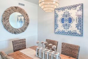 A dining area with a wooden table, wicker chairs, a decorative mirror, a chandelier, and a blue and white patterned wall hanging. - Gulf front Bliss at The Palms 1015B - Orange Beach, AL - Signature Properties (Orange Beach)