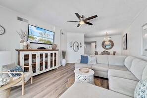 A modern living room with a flat-screen TV, a ceiling fan, and a large grey sofa. - Gulf front Bliss at The Palms 1015B - Orange Beach, AL - Signature Properties (Orange Beach)